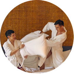Two Asian men in bathrobes pillow fight each other in bed