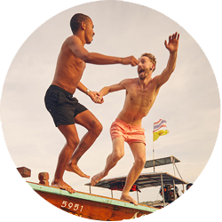 Two men, one in blue swim-shorts and one in yellow swim-shorts, hold hands and jump into the sea from a Thai long boat