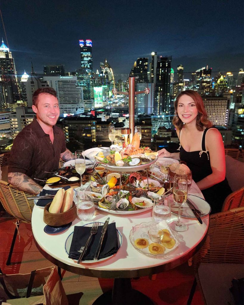 A queer couple sit at either side of a table with a platter of Mediterranean food. The cityscape can be seen in the backdrop. Pastel is a must-visit for those spending one day in Bangkok.