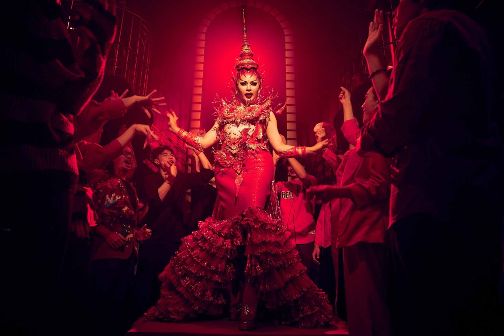 A drag queen in traditional Thai costume, lit up in red, performs for a queer crowd at a Thai gay bar