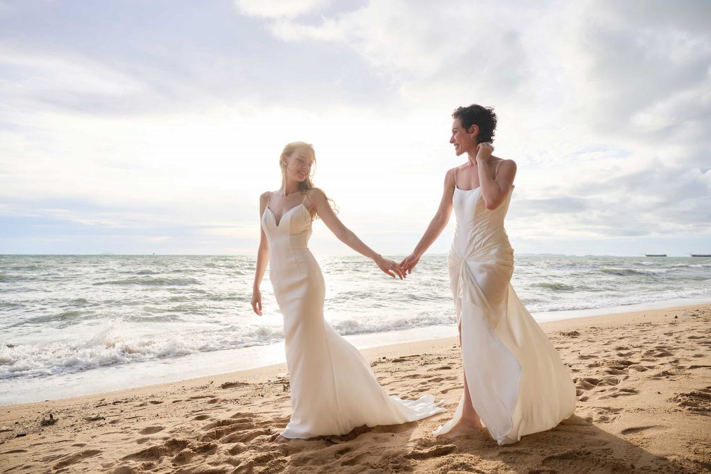 Two women in wedding dresses walk hand in hand down a beach in Thailand