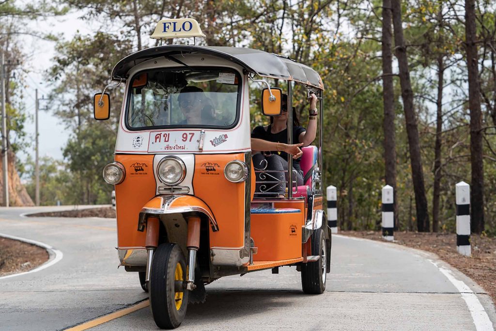 An orange and white tuk tuk driving down a road.