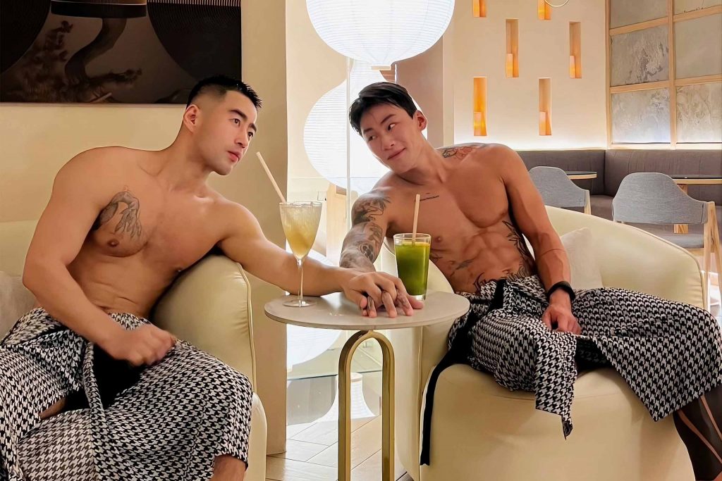 Two men sitting and holding hands at Zen Onsen. They are wearing towels on their waists and have smoothies on the table.