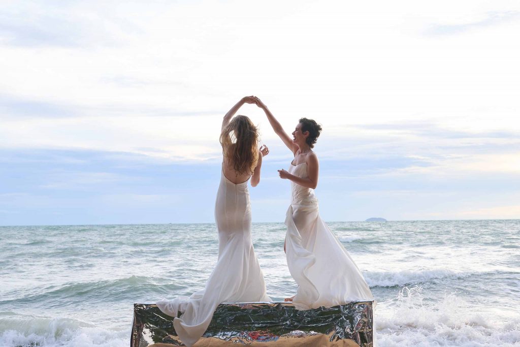 A lesbian couple of newlyweds in their bridal gowns dance in front of the ocean