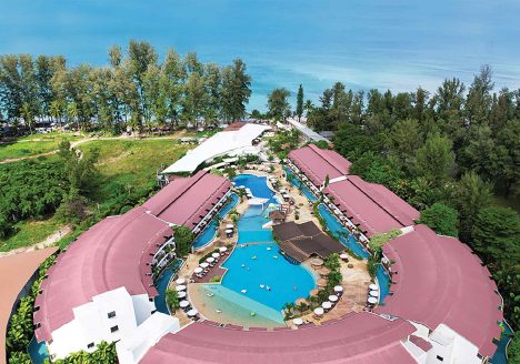 A horseshoe-shaped resort from above. A pool sits at the centre, and the coast is in the distance.