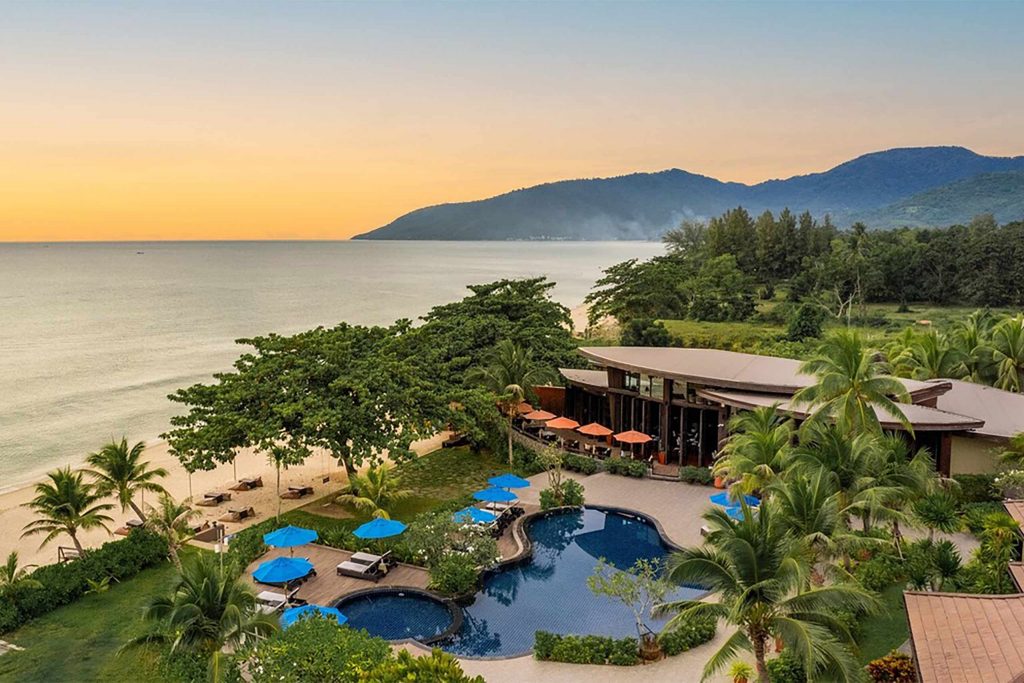 A resort pressed against the ocean at sunset. Silhouettes of hills are in the distance.