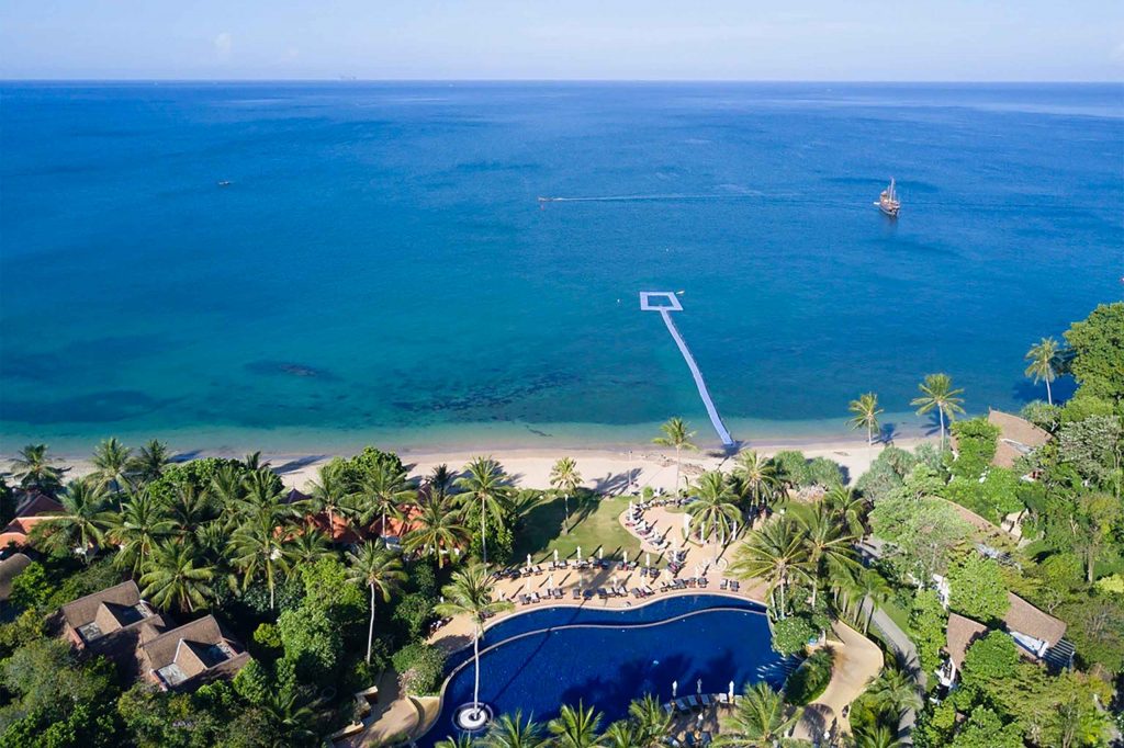 A resort with a pool at its centre pressed against a beach. The wide ocean can be seen, and it stretches out to the horizon.