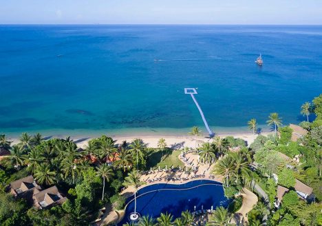 A resort with a pool at its centre pressed against a beach. The wide ocean can be seen, and it stretches out to the horizon.
