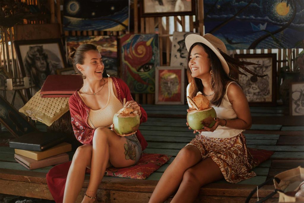 Two lesbian travellers holding coconuts and laughing during Pride Month in Thailand. 