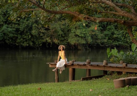 A female traveller sat by a river, surrounded by trees.