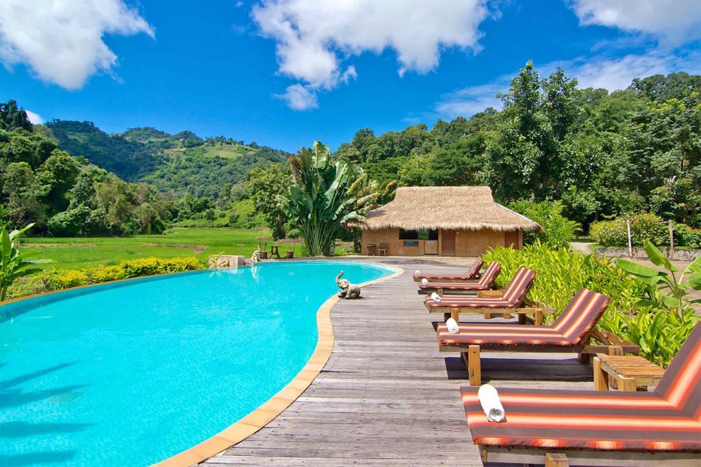 A curvy pool with wooden sun loungers beside it. Lush green hills surround.