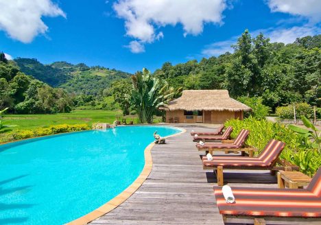 A curvy pool with wooden sun loungers beside it. Lush green hills surround.