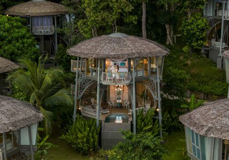 Charming stilted villas standing tall in a forest at twilight with their interiors lit by lanterns.