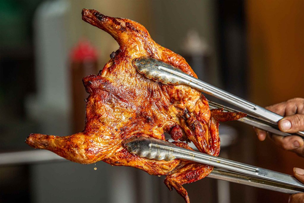 A whole grilled chicken held with metal tongs, its skin golden, crisp and lightly charred.