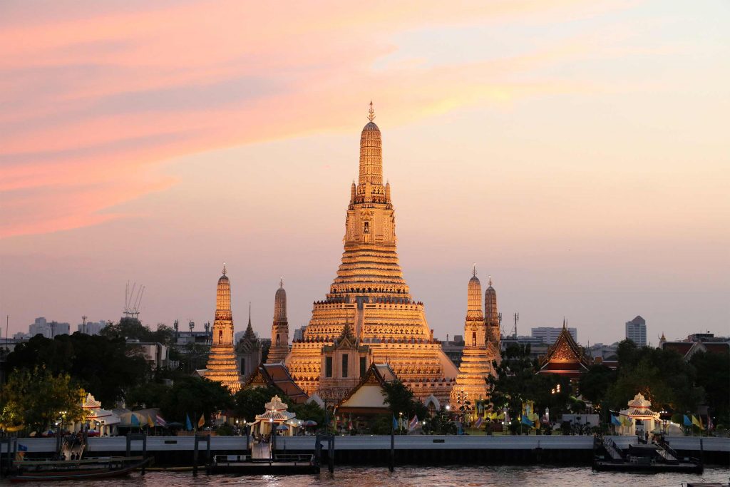 Wat Arun glowing at sunset beside Bangkok's Chao Phraya River, with its ornate central spire dominating the skyline.