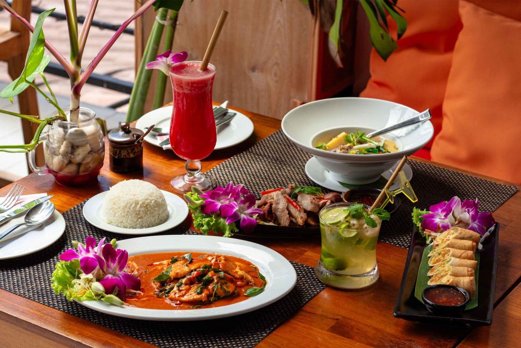 A colourful spread of Thai dishes, rice and drinks served on a wooden table.